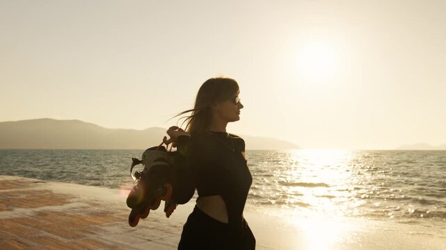Young woman in black crop top carrying roller skates walks along beach at glowing golden sunset. Lens flare, sparkling sea, mountain silhouettes. Active summer freedom and outdoor lifestyle.
