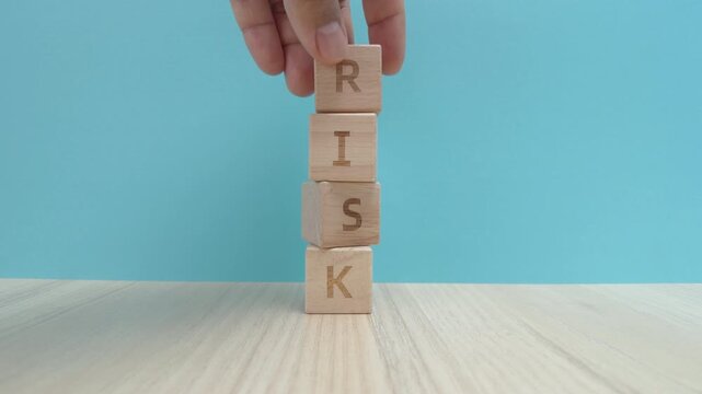Hand places a wooden block to complete the word RISK on a stack that then collapses on a table. Concept of business failure, financial instability, and danger
