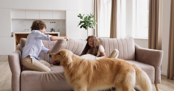 Children siblings holding pillows, having fun, fighting, playing together on sofa in living room at home, playful golden retriever dog nearby. Family bonding, weekend activity, childhood indoors games