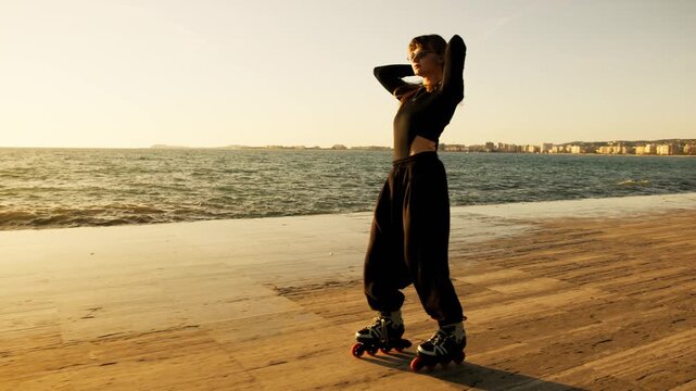 Full length young woman in black skating toward camera on golden seafront at sunset. Wind in hair, coastal city and sea on horizon, long shadow on glowing deck. Active summer freedom and lifestyle.
