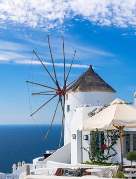 Vista vertical de un molino de viento tradicional en Santorini, Grecia, destacando la arquitectura cicl&aacute;dica blanca contra el azul profundo del mar y el cielo con nubes.