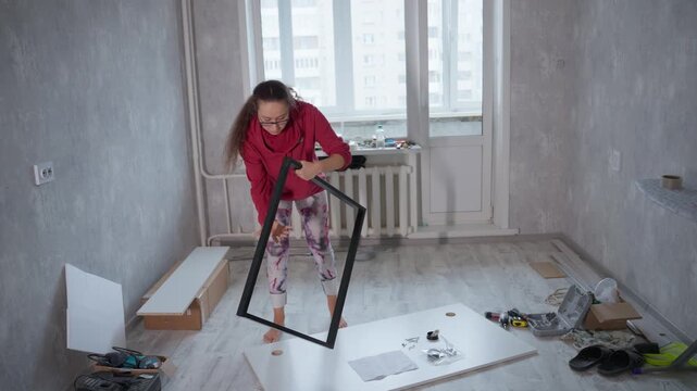 Young Caucasian Woman Assembling Furniture Frame In Empty Apartment, New Homeowner Inspects Black Metal Leg Frame, Scattered Tools, Instruction Sheet On Floor, Barefoot Casual Hoodie, Soft Daylight