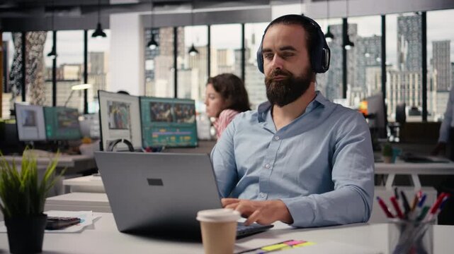 Headphones in office, working with focus music, business employee at desk, multitasking for productivity. Adult man sitting in modern office environment. Listening to audio and solving tasks.