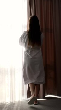 Young woman opening curtains and letting sunlight into room. Fresh morning start, lifestyle, wellness and positive energy at home or hotel.