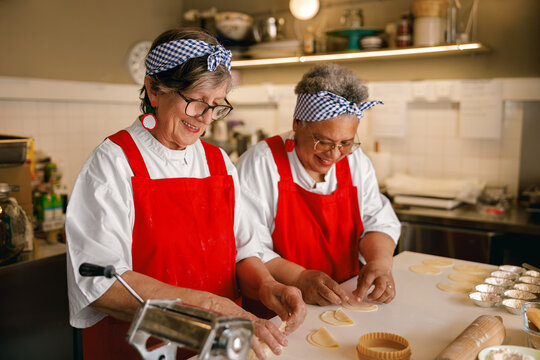 Two diverse senior women, genuinely smiling, collaborate on shaping dough for traditional food in a professional kitchen, bathed in natural soft light.
