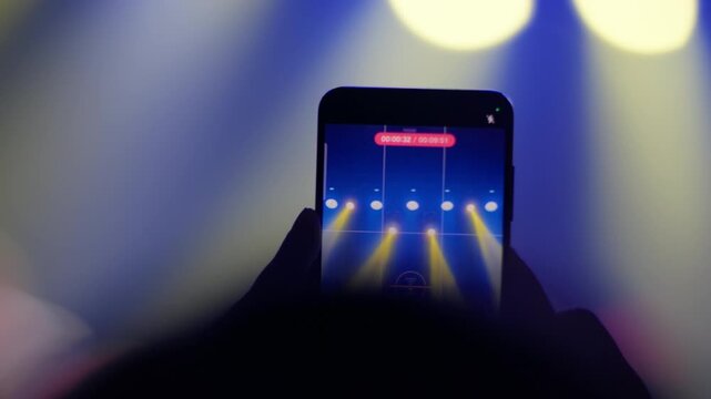 Hand holding smartphone recording concert stage with yellow and blue spotlight beam, video recording timer visible on screen, slow motion