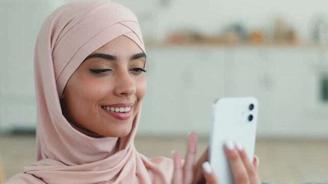 Happy smiling Arabian Muslim woman in hijab using mobile phone scrolling at home positive female Islamic girl holding smartphonegadget addict social media virtual communicate online technology indoor