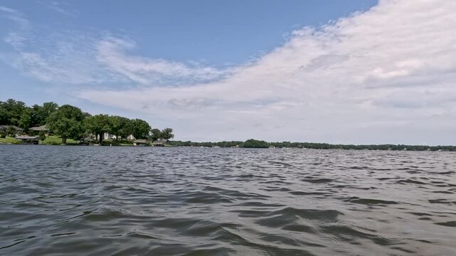 Smooth POV gliding across blue water toward a lush green shoreline and waterfront homes. Vivid greenery and bright summer sunlight highlight a Midwest nautical lifestyle.