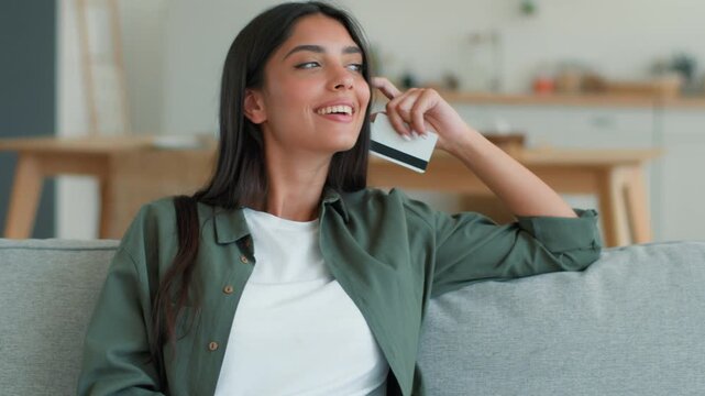 Happy joyful Indian Muslim woman using mobile phone online shopping at home couch kitchen ordering food delivery entering credit bank card Arabian female girl client customer buyer purchase indoors