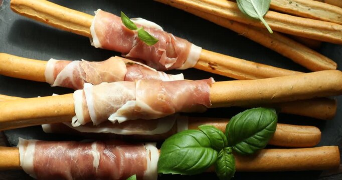Grissini breadsticks with prosciutto and basil on wooden table, spinning. Top view