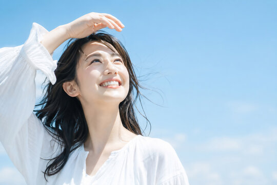 青空の下で眩しそうに目を細め笑顔を浮かべる若い女性 紫外線対策やスキンケアのイメージ Generative AI