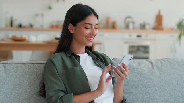 Happy smiling Arabian Indian woman using smartphone at home indoor chatting browsing social media shopping online Hispanic female girl holding mobile phone internet addict app net web cellphone device