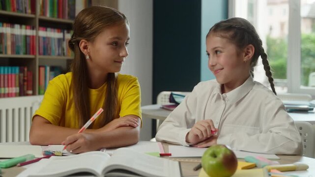 Cute Caucasian little girls children reading book together desk in classroom schoolgirls students assisting helping friend kids classmates learning lesson help writing task education teamwork school