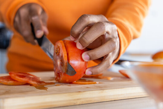 African American man wearing orange shirt slicing ripe tomato on wooden board with chef knife