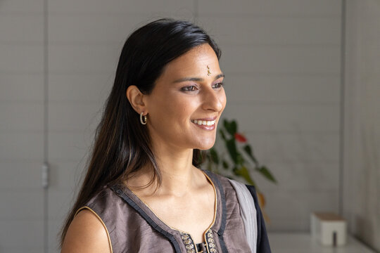 Asian female smiling and looking right in room, wearing embroidered tunic, bindi and gold hoops