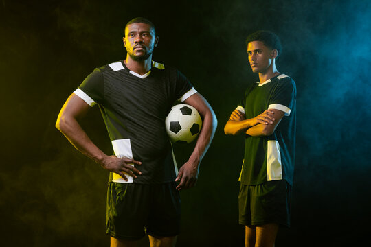 Male teammates posing in studio with smoke, holding soccer ball, wearing black jerseys and shorts