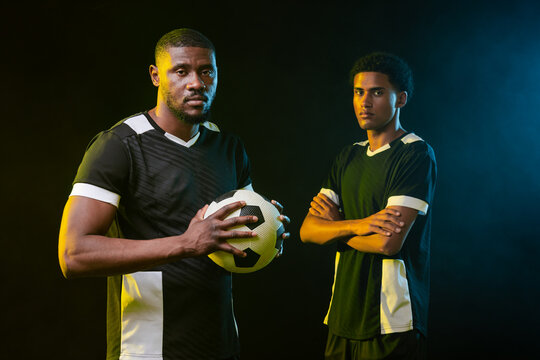Male soccer teammates posing in studio under rim lighting, one holding b and w ball in black kits