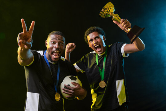 African male teammates celebrating in studio wearing jerseys and medals, holding ball and trophy