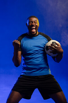 African male holding soccer ball shouting, celebrating in studio with haze blue jersey black shorts