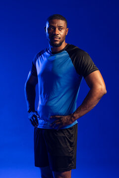 Mid-30s man posing with hands on hips on deep blue backdrop, wearing blue athletic shirt
