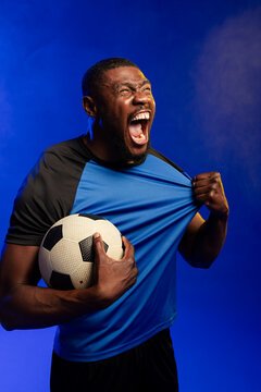 Mid adult African man shouting while holding soccer ball and pulling blue-black kit, studio haze