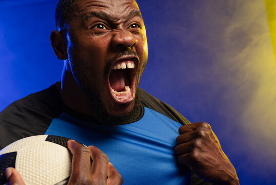 Male footballer shouting holding textured black-and-white ball pulling blue-black jersey in studio