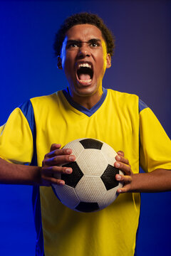 Adult African male shouting gripping b and w ball in yellow-blue jersey blue-yellow rim purple backd
