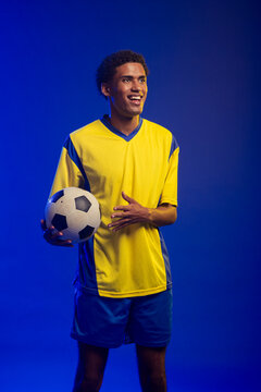 African American man holding black-and-white soccer ball, wearing yellow-blue kit in studio