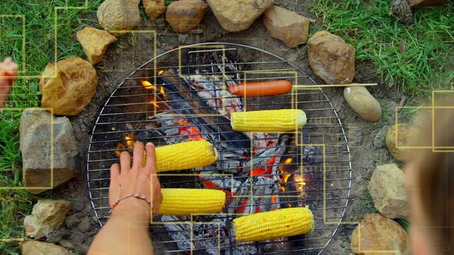 Embers flaring, hands turning corn and moving skewered hot dog to control charring for sharing