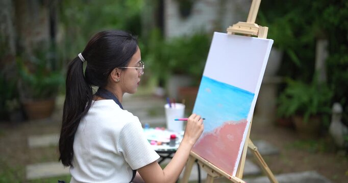 asian woman painting outdoor landscape with focused expression during relaxing session sitting in garden using brush to blend tones on canvas enjoying peaceful hobby moment surrounded by greenery