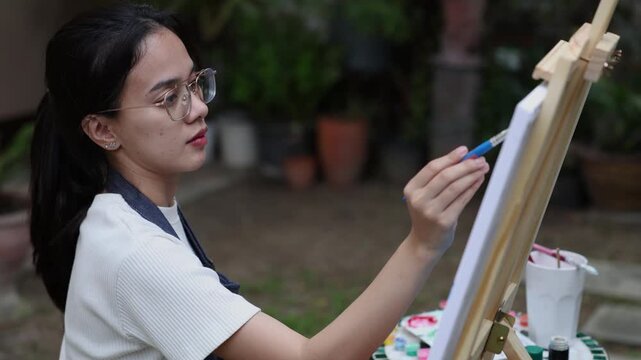 asian female artist creating soft background on canvas using blue brushstroke in relaxing garden setting with focus enjoying peaceful moment of creativity and expression during outdoor painting 