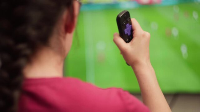 Woman holding a remote control watching a soccer match on television at home, enjoying her leisure time while supporting her favorite team during a live championship
