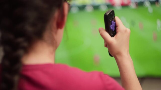 Woman watching a soccer game on a streaming television service and holding a remote control, with a focus on her hand and the device while switching channels at home