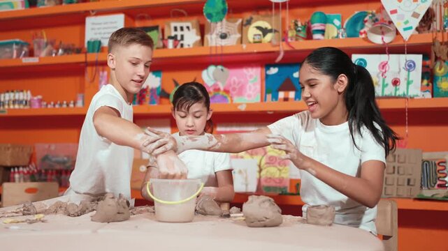Multicultural happy student playing by throw clay while modeling cup in pottery class. Caucasian high school girl pointing at boy while smiling smart boy laughing at art lesson. Education. Edification