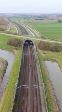 Vertical screen Aerial View Of Rail Tunnel Exit Leading Into Straight Double Tracks Across Farmland, Overhead Lines Visible, Calm Rural Atmosphere And Narrow Water Channels.