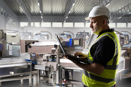 Industrial production management, engineer operates machinery with laptop, monitors manufacturing line, equipment performance and factory process optimization