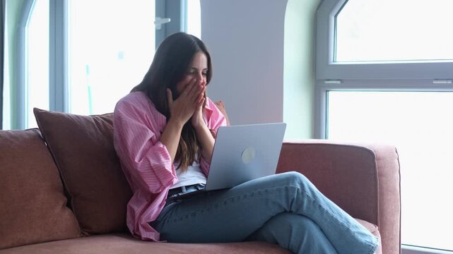 A happy young woman sits comfortably on a sofa, reacting with excitement and joy to something seen on her laptop screen, celebrating a pleasant surprise or achievement in her cozy living room.