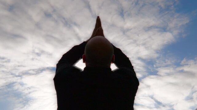 View of a bald man from behind, showing upper body, with hands and arms up, praying to the heaven above as his palms are together with a cloudy sky in the background.