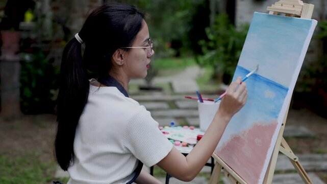 young asian woman artist holding blue brush painting canvas while sitting outside surrounded by nature focusing on calm creative process during quiet moment in backyard garden