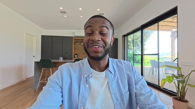 A happy young Black man smiles genuinely at the camera, possibly on a video call from his bright, modern home. His friendly expression conveys joy and warmth.