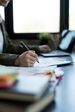 Close-up of businesswoman hand signing a contract or writing on a document while working at an office desk with a laptop.