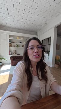 A cheerful young woman in glasses smiles and gestures while talking to the camera, possibly on a video call or recording a vlog. She conveys a friendly and positive vibe from her modern home office.