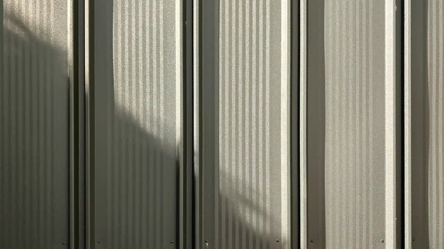 Corrugated metal wall texture with moving shadows, silver industrial siding background, metallic surface with rhythmic patterns, candid style video, architectural exterior detail, light and shadow.