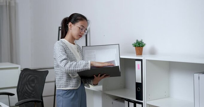 Asian female opening file folder and flipping through documents while standing near white cabinet in tidy home office reviewing paperwork with focus and professionalism in quiet moment