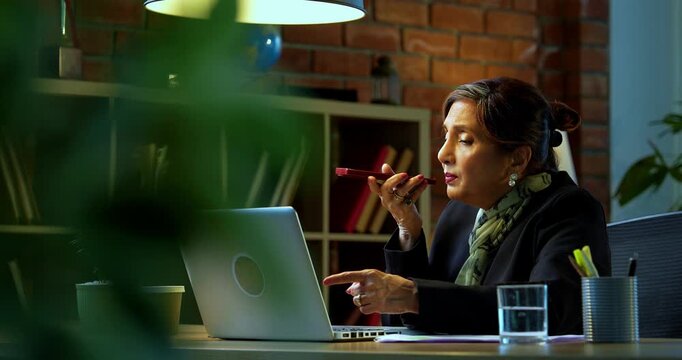 Senior Indian businesswoman in corporate attire attending important phone call on speaker phone via smartphone at workstation with laptop in cozy office space experienced female executive desk