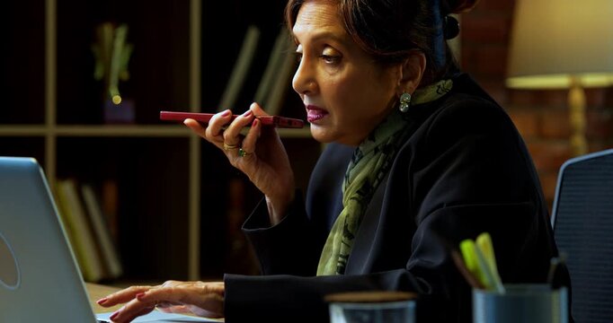 Senior Indian businesswoman in corporate attire attending important phone call on speaker phone and typing on laptop in modern cozy office space experienced female executive with white hair