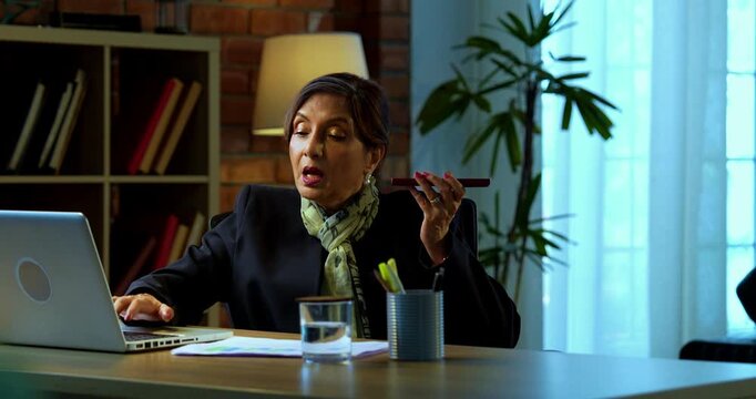 Senior Indian businesswoman in corporate attire attending important phone call on speaker phone and typing on laptop in modern cozy office space experienced female executive with white hair