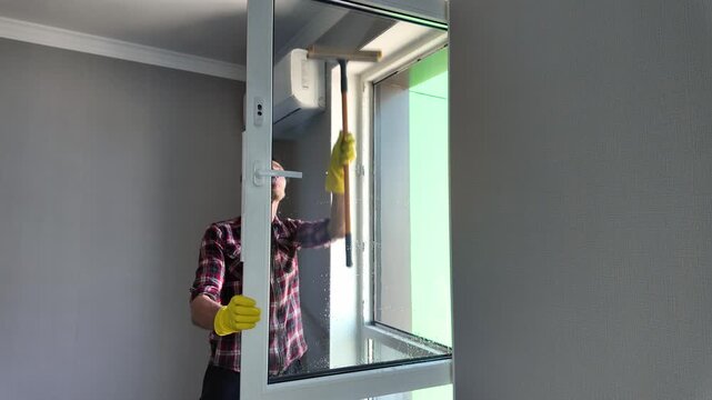 Caucasian man in yellow rubber gloves diligently cleaning a large window pane with a professional squeegee during household chores