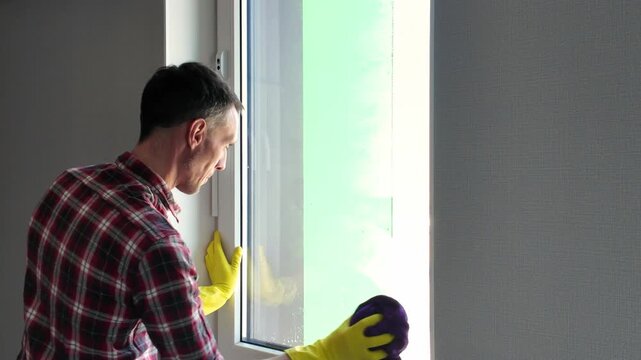 Man in a plaid shirt and yellow rubber gloves diligently cleaning a house window with a purple cloth. Professional household chore concept