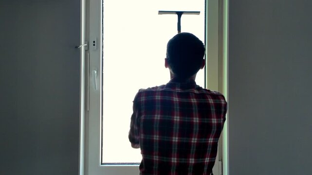 A man cleaning a window with a squeegee in a modern apartment, rear view